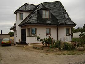 Nietypowy, parterowy dom z poddaszem użytkowym, o zaokrąglonych narożnikach i ciemnym dachu z dachówki ceramicznej, widoczny z perspektywy ogrodu, z żółtym samochodem zaparkowanym obok.